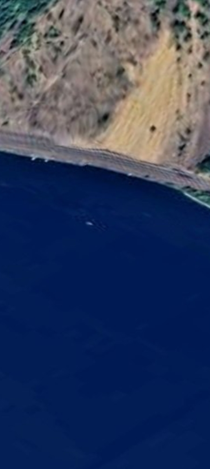 Mid-zoom satellite view of the Columbia River shoreline along Highway 14 Washington side showing the bottom edge of the yellow mountainside exposure meeting the highway shoulder and the water below with subtle wake patterns and surface disturbance visible in the dark blue river water immediately offshore at the same coordinate where the USO is documented in the subsequent tighter frame