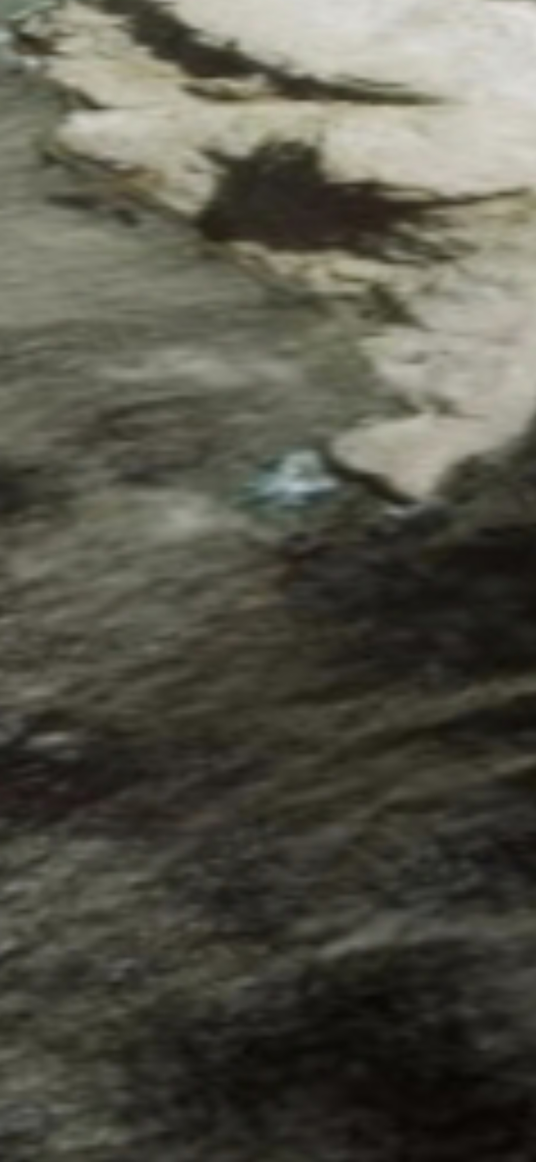 Close-up zoom of the small bright reflective fly-in craft resting on the rocky surface of Isla Cardones just off Mazatlán Bay with the dark surrounding water visible and the metallic surface of the craft showing distinctly against the natural rock terrain