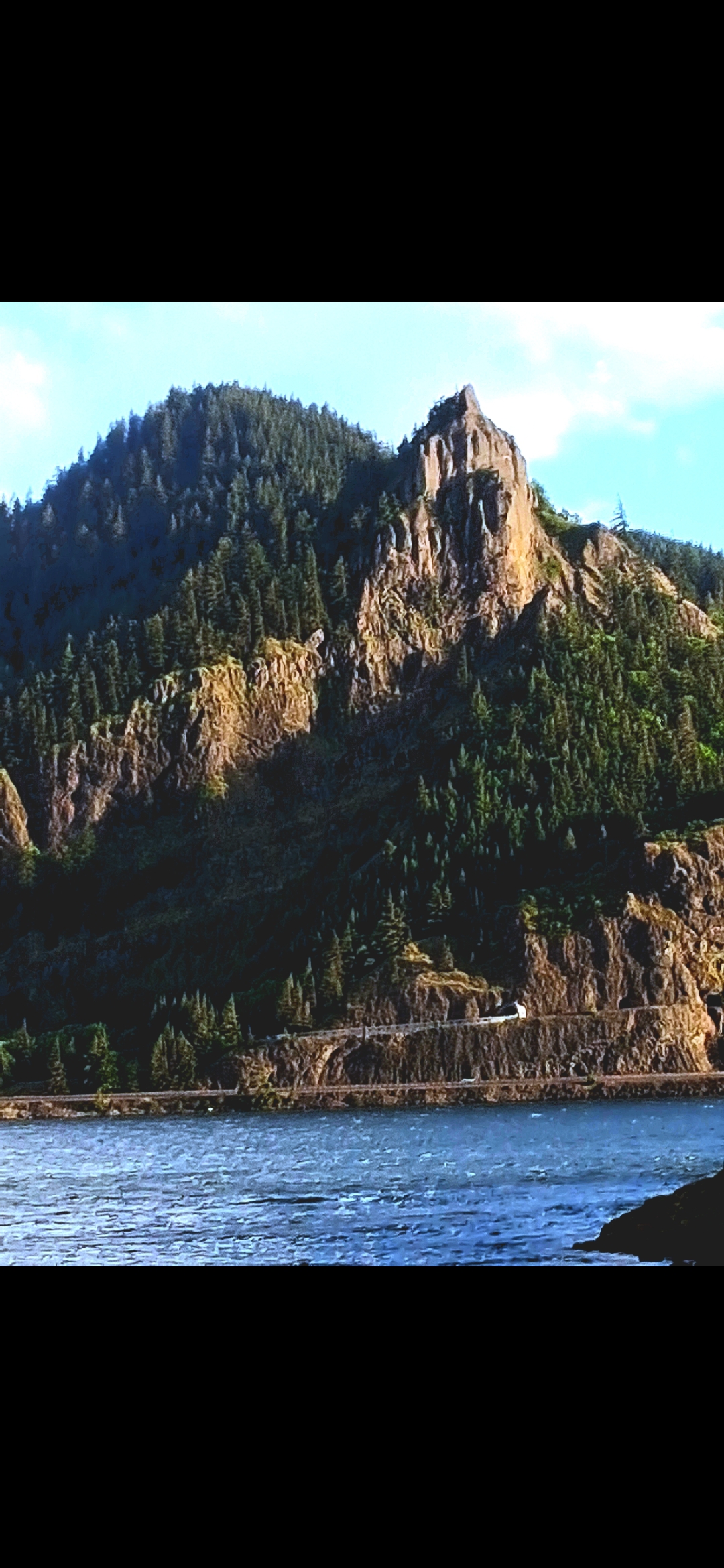Wide daylight photograph of Mitchell Point Oregon taken by the Perceiver from across the Columbia River on the Washington State side showing the prominent basalt headland rising above Interstate 84 along the south bank with the river in the foreground and the wooded ridge line behind the formation establishing the formation in its full landscape context