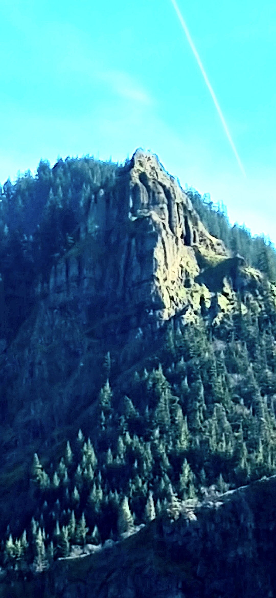 Closer daylight photograph of Mitchell Point Oregon basalt headland with the rocky outcrop in the upper center of the frame and the face geometry beginning to resolve in the wall of the cliff a brief sun-flare streak visible in the upper right corner of the sky