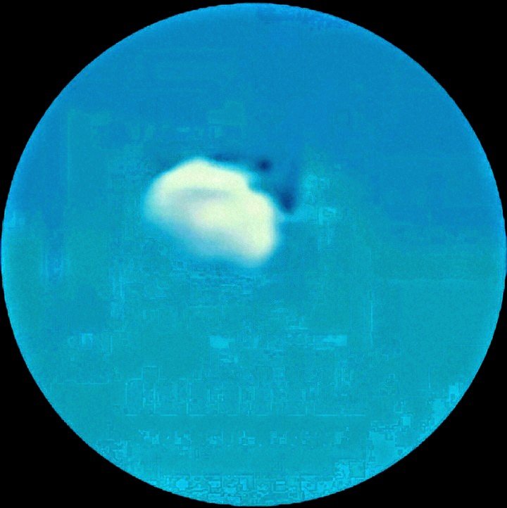 Third moon imposter capture shown as a cyan tinted circular zoom crop with a luminous white pale core embedded within a cloud formation and scattered small dark discrete particle shapes dispersed across the frame in the characteristic drop pattern