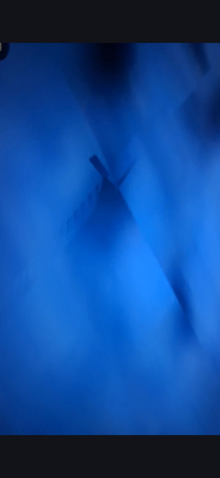 Opening frame of the Pacific underwater installation capture showing a dark cross-shaped or anchor-shaped silhouette embedded within the deep blue camouflaged ocean surface viewed from satellite altitude with infrared frequencies penetrating the camel-layer film overlay