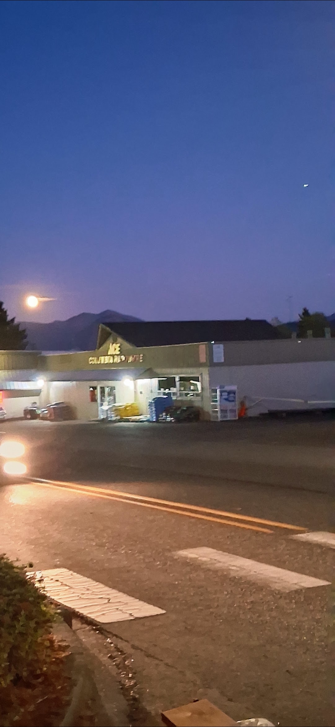 Captura amplia al anochecer del comercio Columbia Ace Hardware en Stevenson Washington al crepúsculo con un objeto rectangular oscuro visible en la parte superior derecha del cielo sobre el estacionamiento y una luna luminosa en el lado opuesto del cuadro con crestas Cascade siluetadas en el fondo