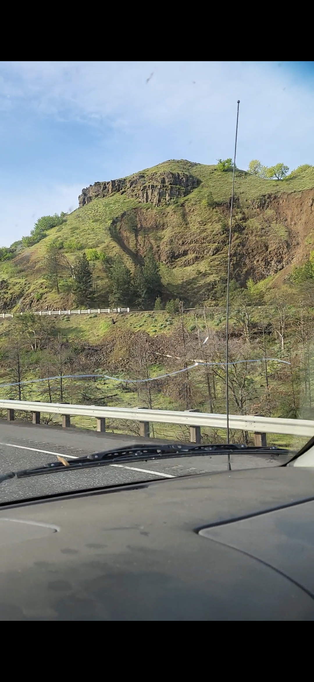 Wide-angle raw unprocessed dash mounted phone capture from the Perceiver vehicle approaching the western entrance of The Dalles Oregon from Hood River along Interstate 84 showing a south facing grass and basalt hillside on the right side of the highway with the carved facial geometry visible at the upper ridge level pending contrast enhanced versions inbound