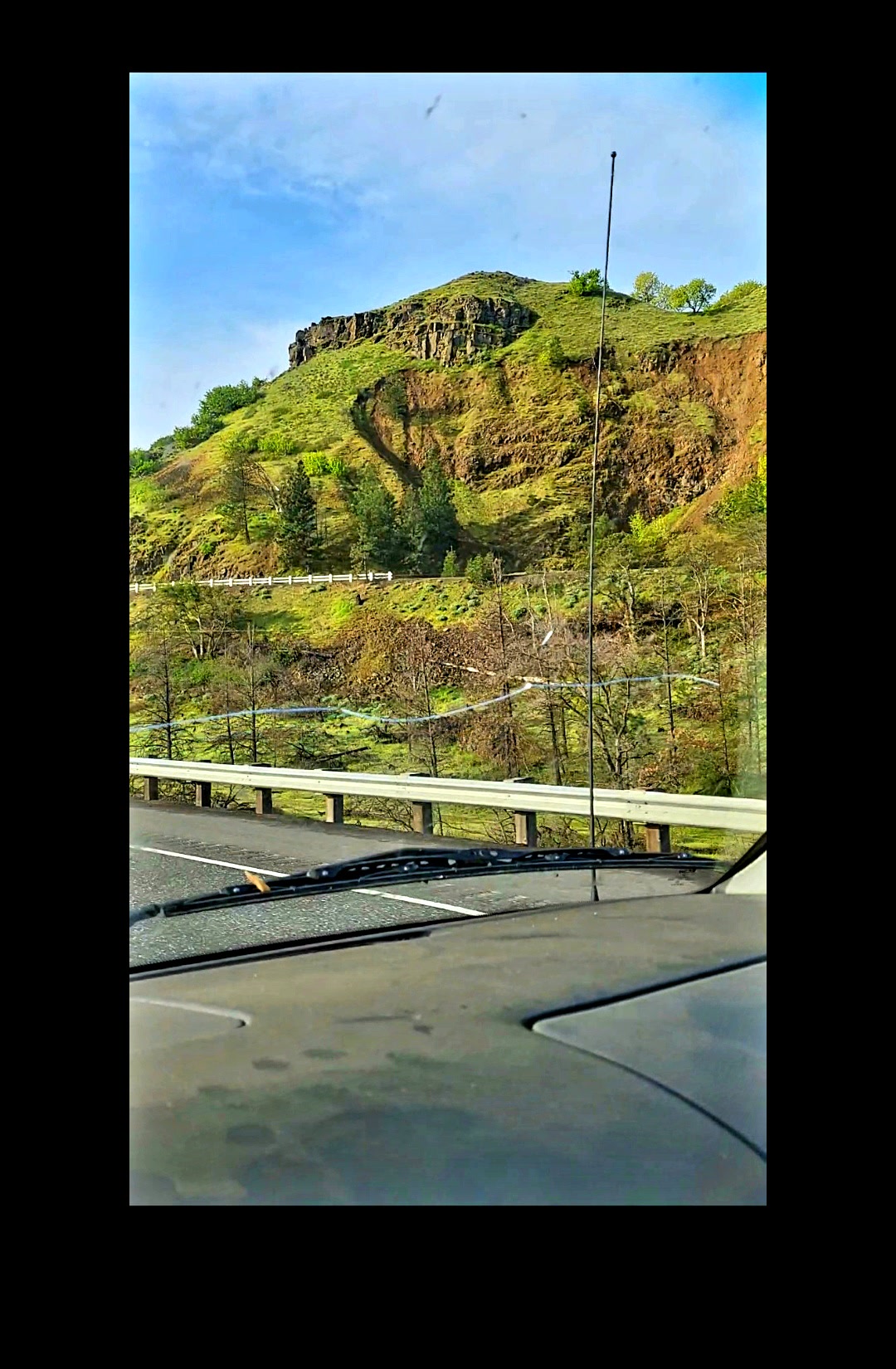 Second raw vantage of the western entrance hillside at The Dalles Oregon dashboard mounted phone capture cleaned of phone gallery UI chrome showing the same carved facial geometry on the upper ridge under a bright blue sky for cross verification of the morphology against the first raw approach frame the eye feature resolves on the upper portion of the carving with an adjacent green monitor sphere observation node to its left